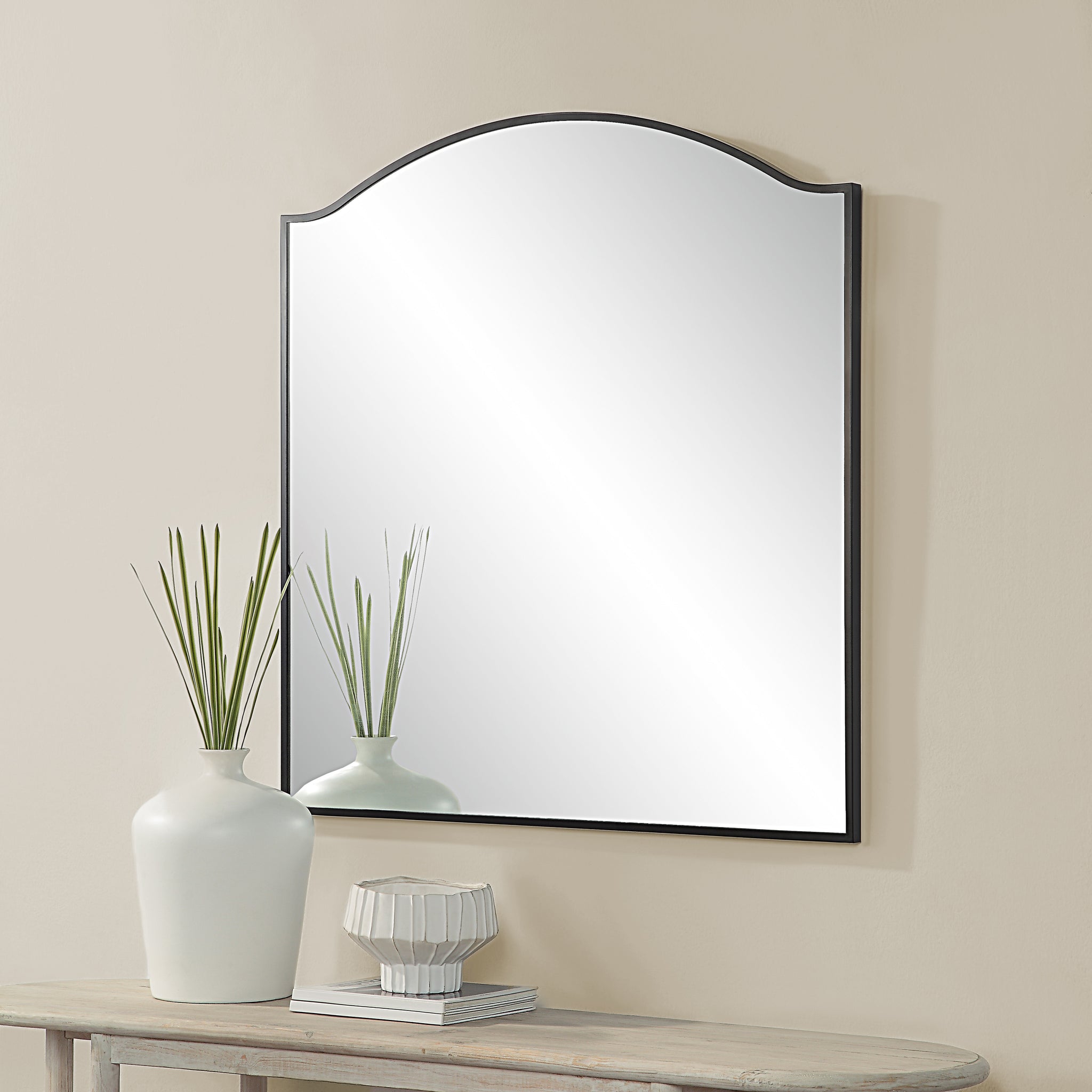 Bandon Wall Mirror displayed above a table with decorative vases, featuring a black frame and curved top.
