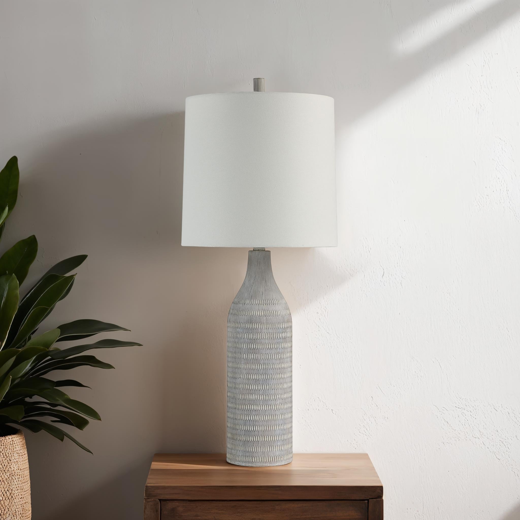 Baracoa table lamp on a wooden table, showcasing its gray base and white shade with plants nearby.