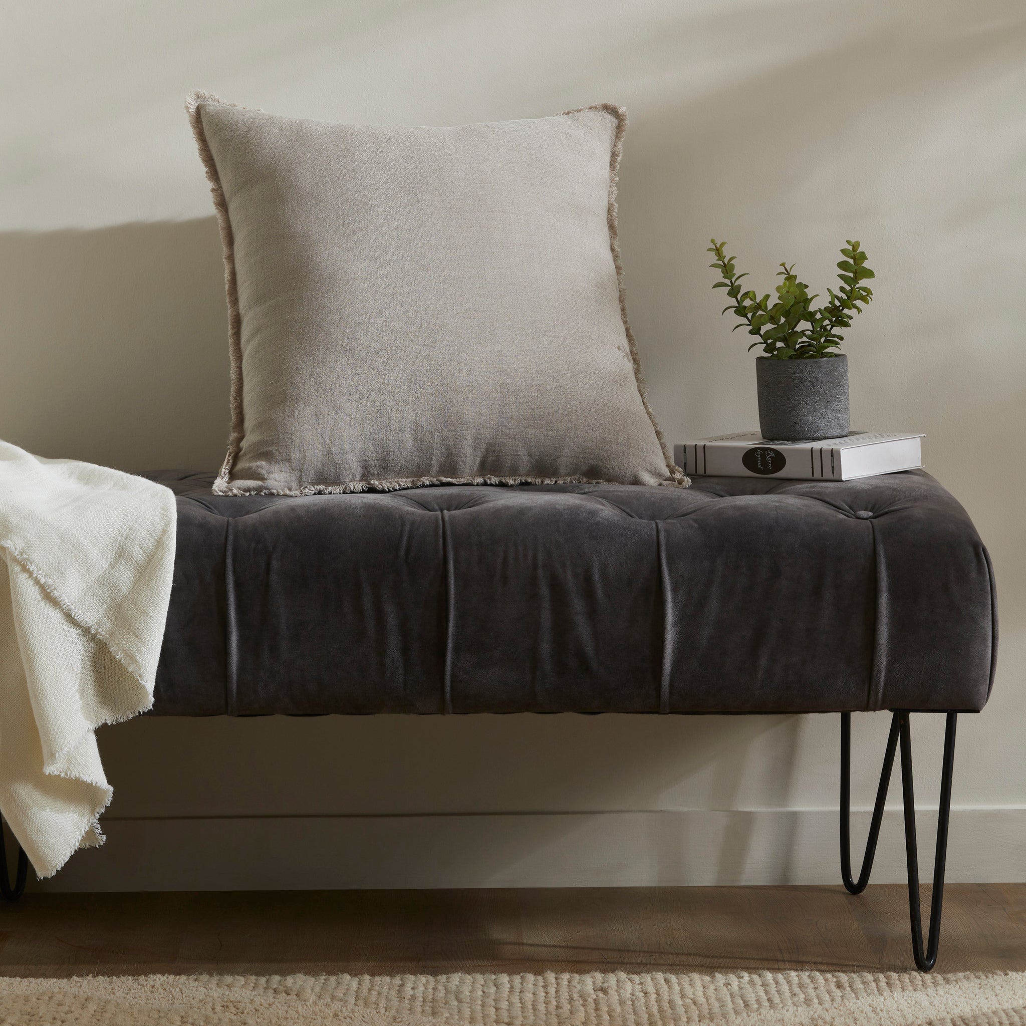 Ignatia Indoor Pillow in light beige placed on a dark tufted bench with a plant nearby.
