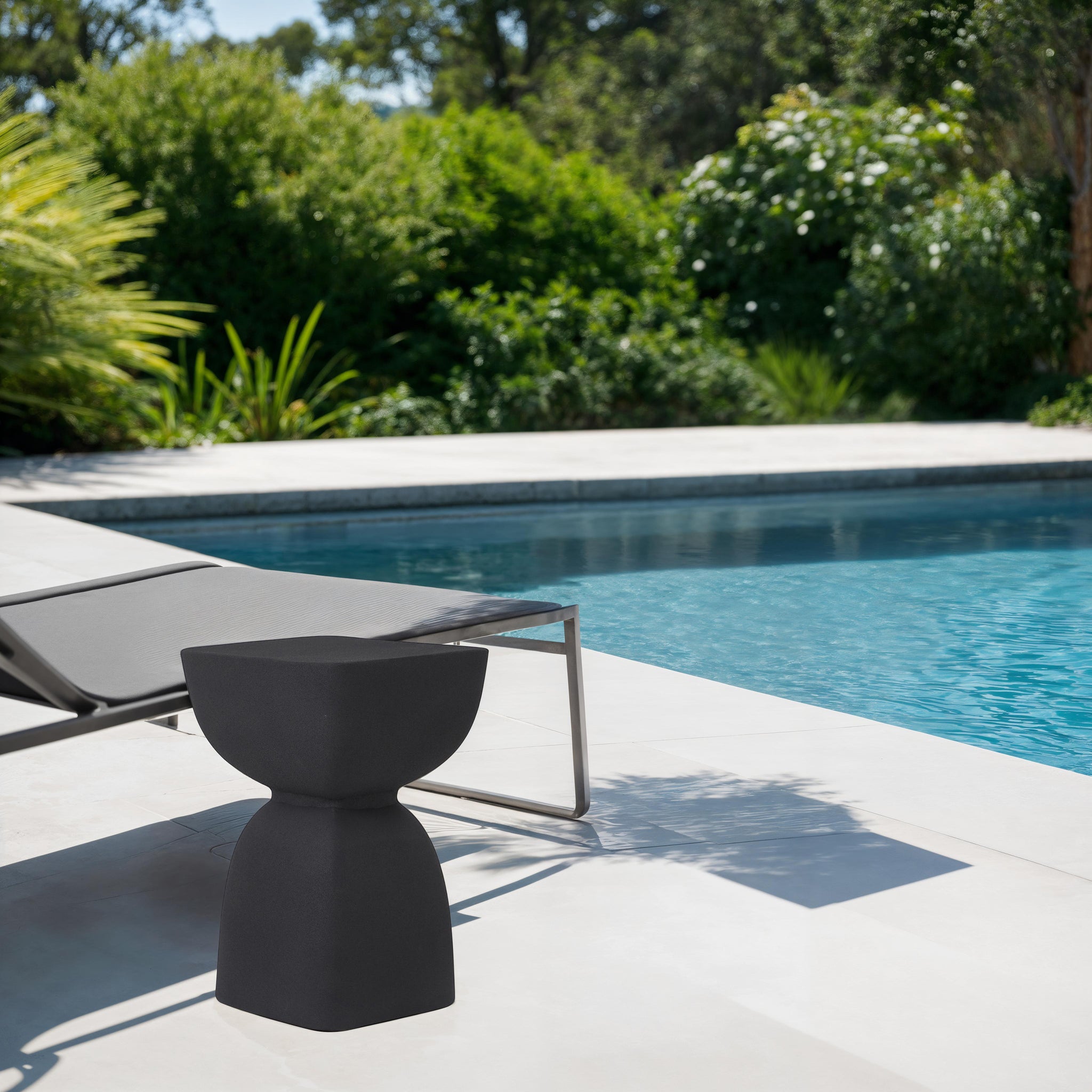 Frans Outdoor Side Table in black beside a pool, surrounded by greenery.