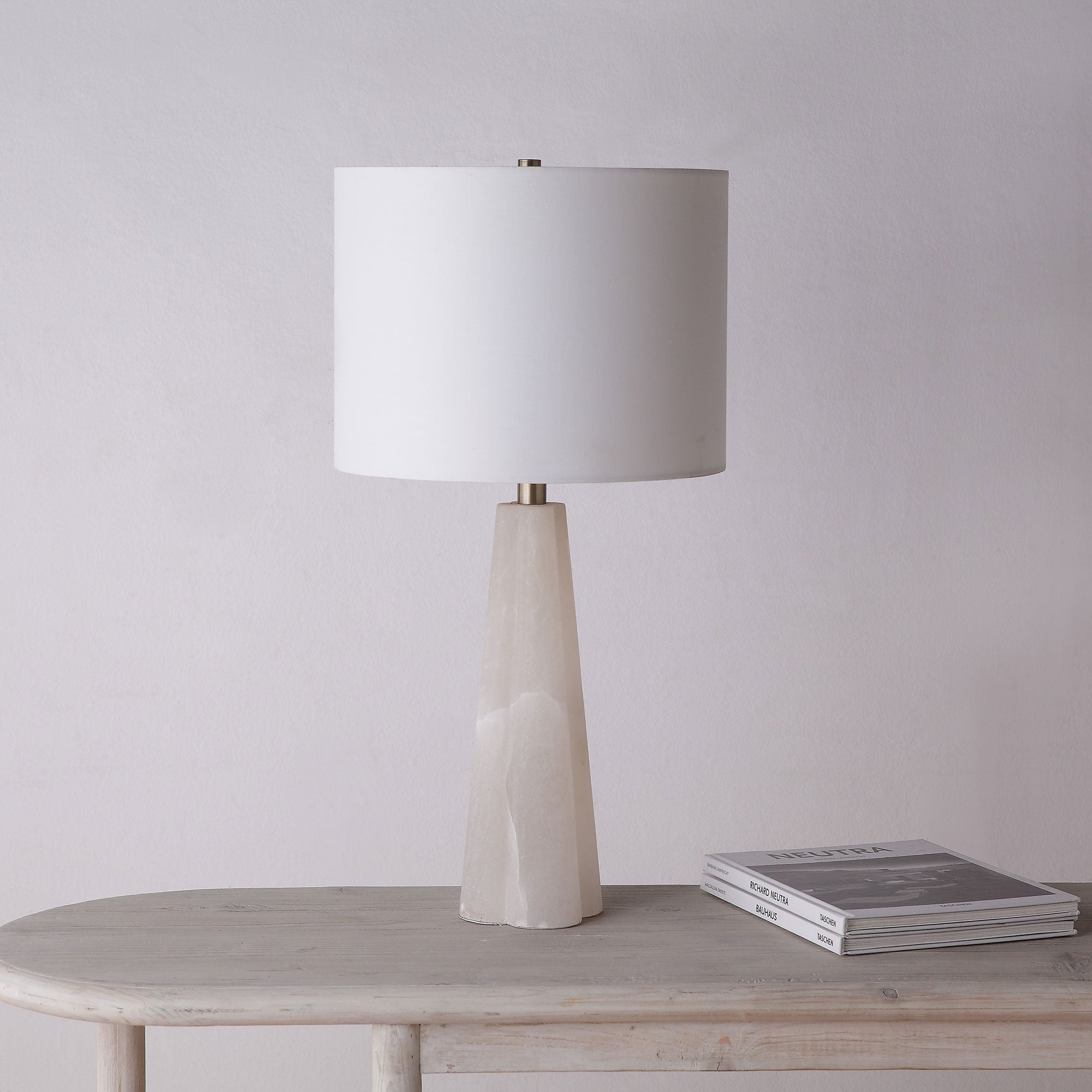 Rima Table Lamp on a wooden table, featuring a white shade and beige stone base against a neutral wall.