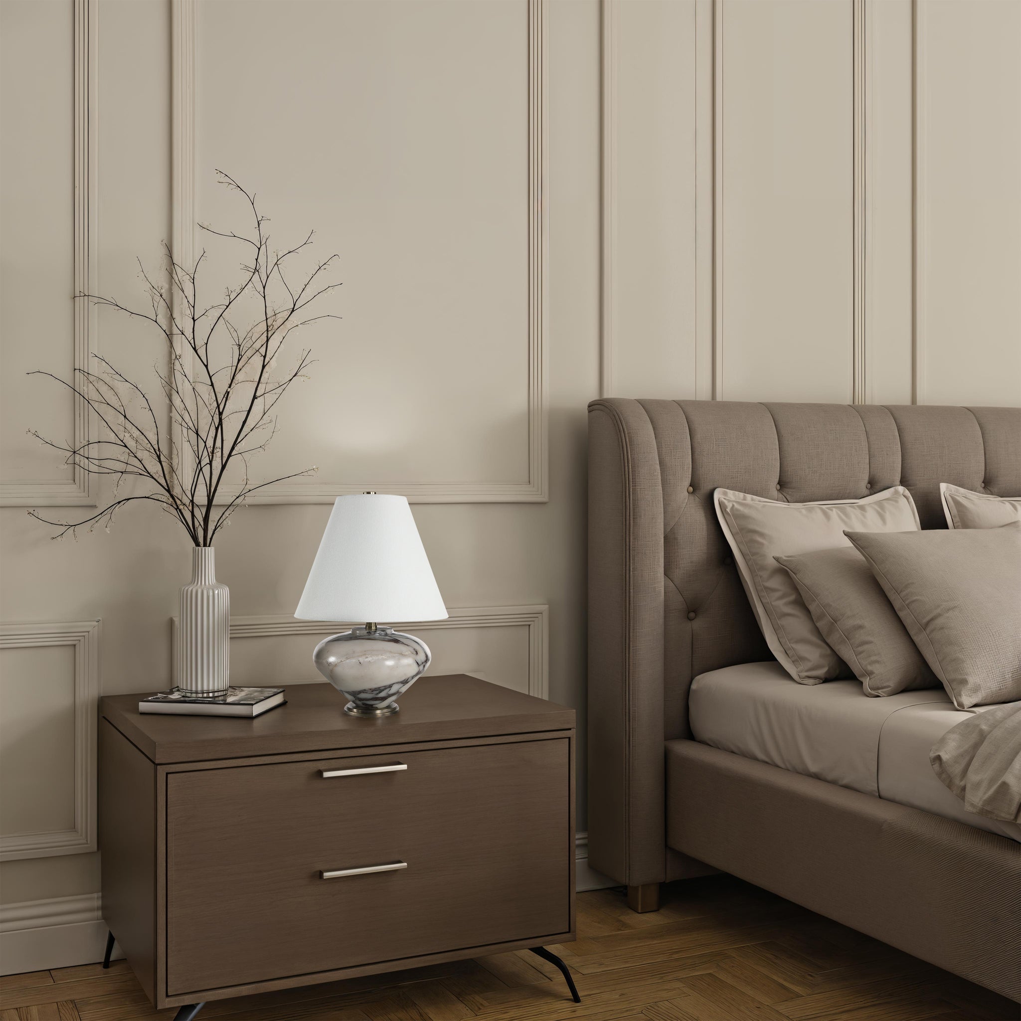 Monumento Table Lamp on a nightstand beside a tufted bed, with decorative pillows and a vase.