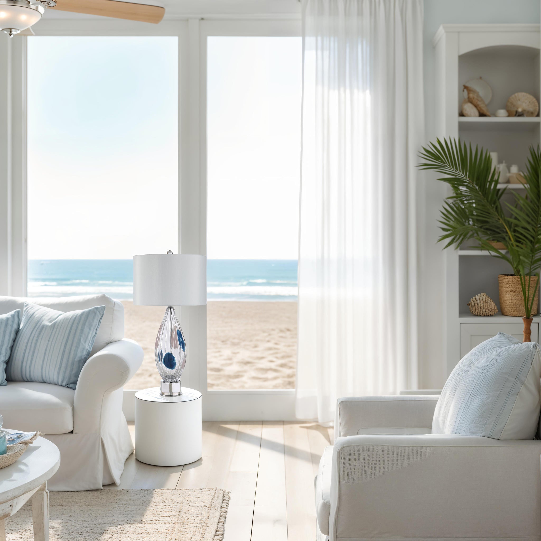 Luxor Table Lamp in a bright living room setting with ocean view, featuring a white shade and glass base
