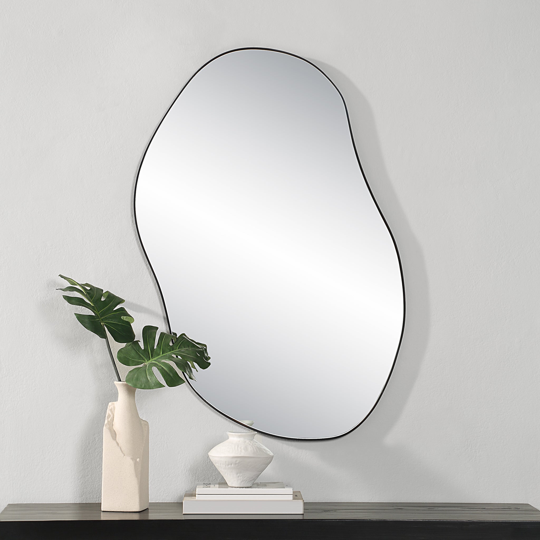 Jane Wall Mirror with an organic shape, featuring a black outline, mounted on a light wall, alongside decorative vases and greenery.