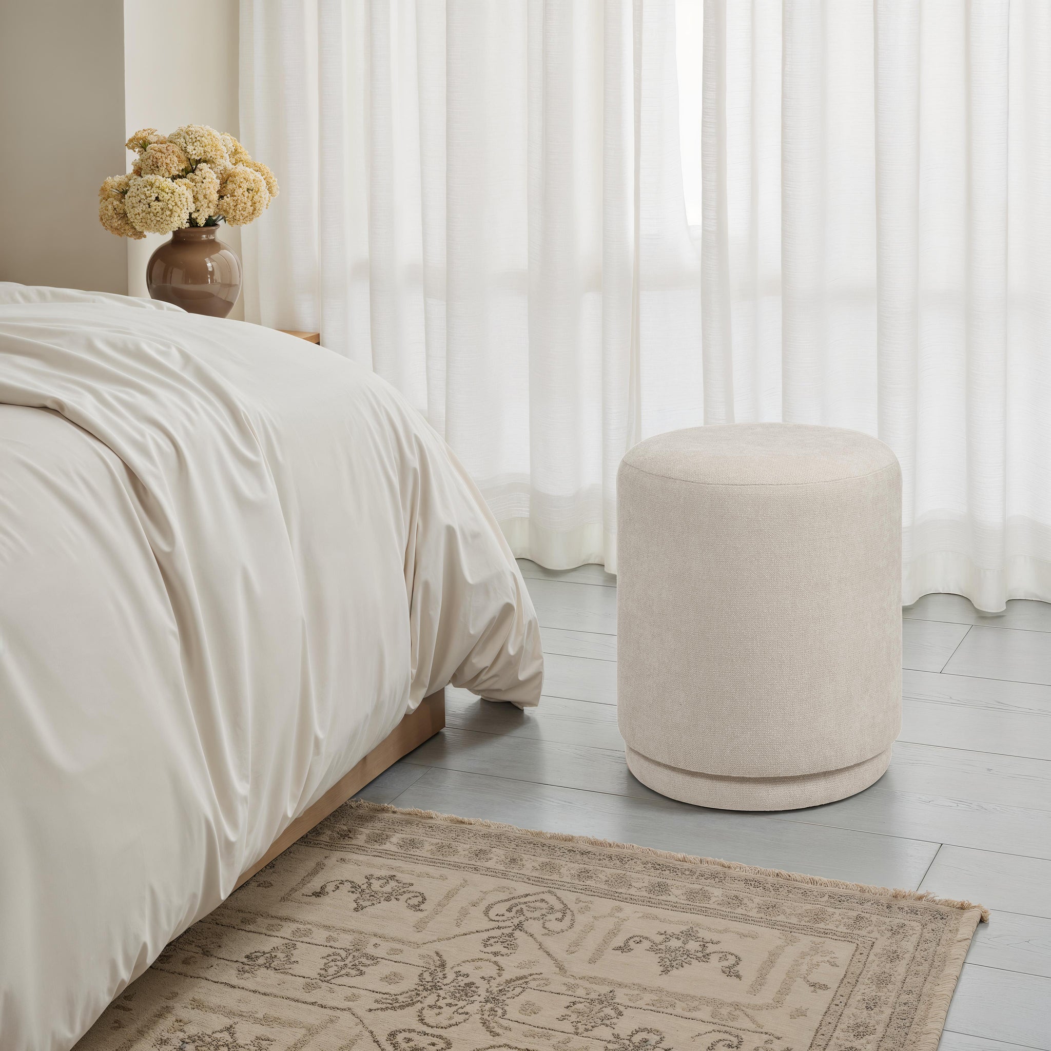 Persea stool placed beside a bed in a softly lit room with curtains and a floral vase.