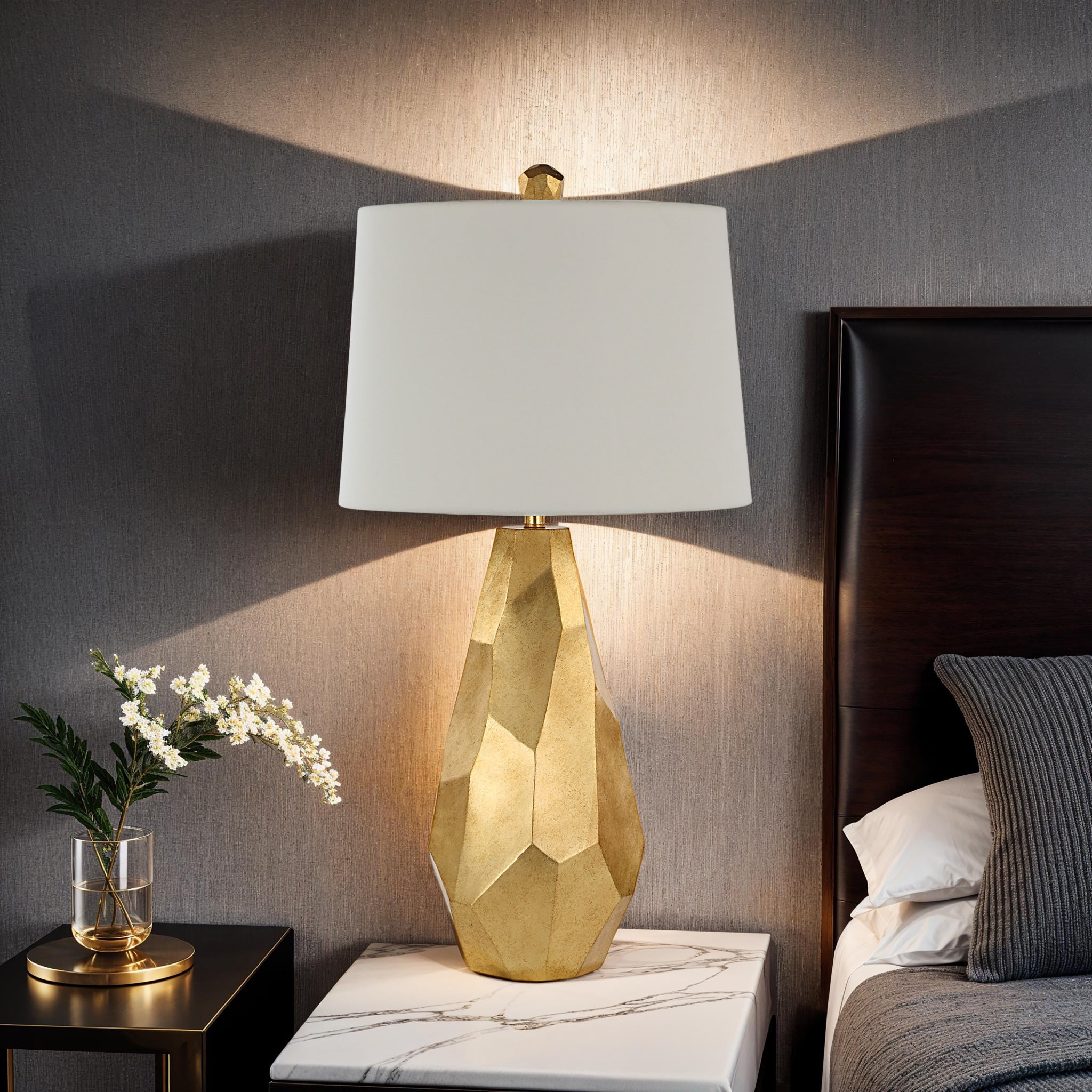 Sanae Table Lamp by Renwil in a bedroom setting, casting light against a textured wall.