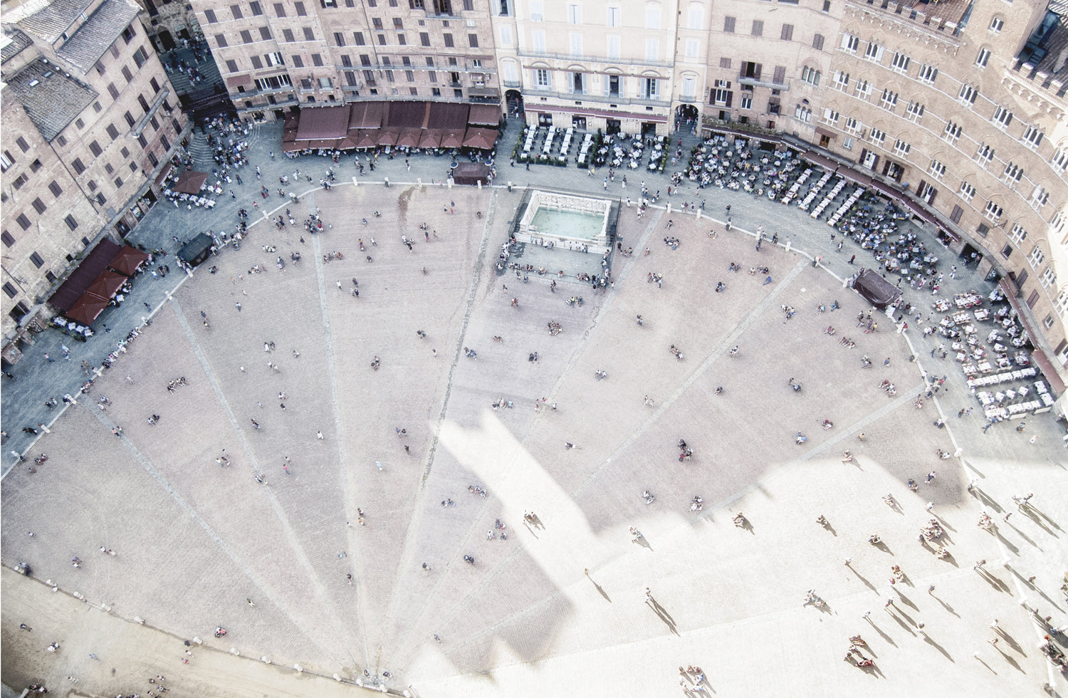 Piazza De Campo, an aerial view of a bustling square with people, cafes, and a central fountain, featuring sandy tones.