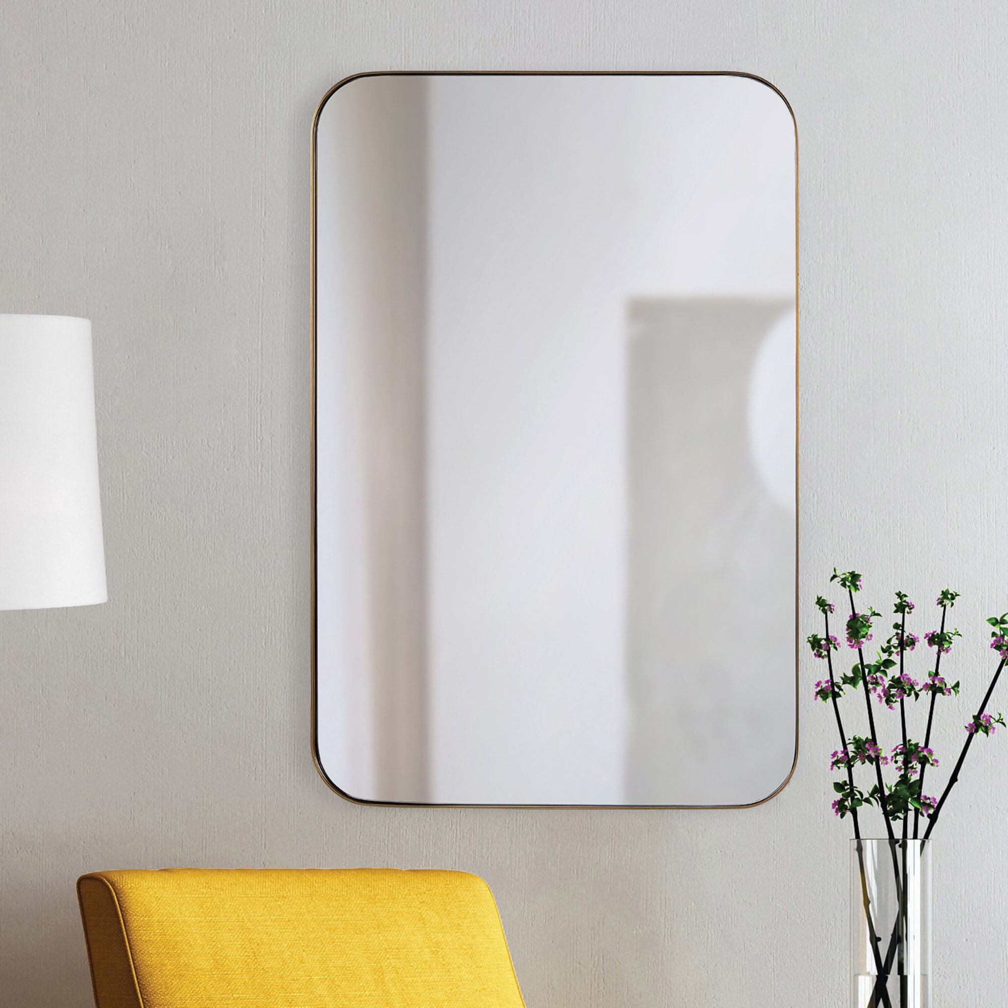 Edwin mirror mounted on a wall beside a yellow chair and a vase with flowers.