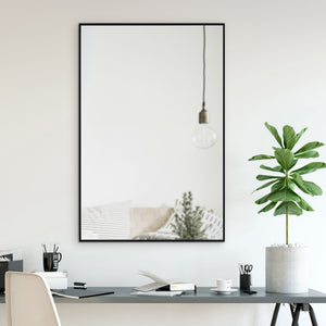 Reynolds mirror with a black frame reflecting a bright room, featuring a hanging light and a potted plant.