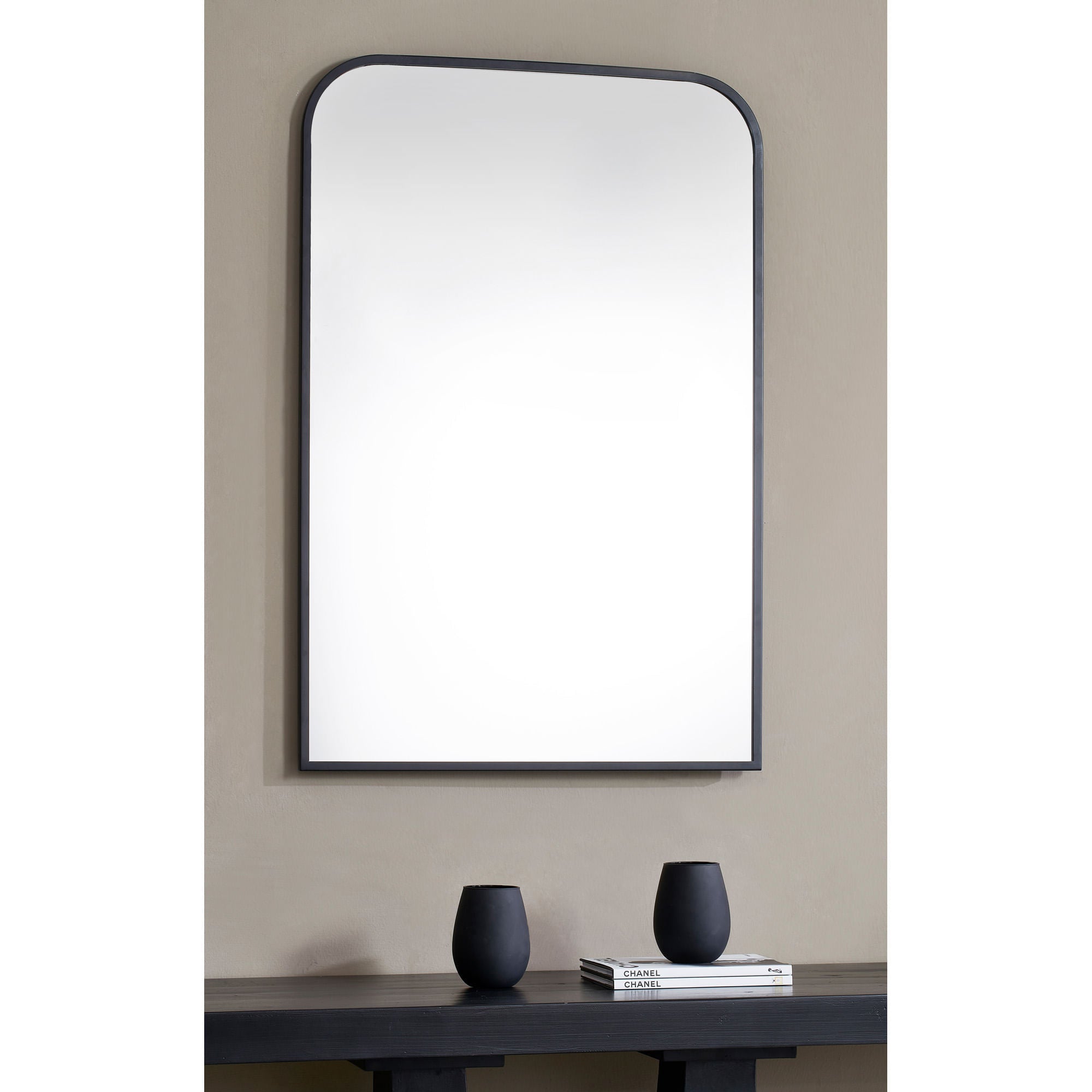 Jackline mirror with a sleek black frame, featuring a rounded top, mounted on a beige wall above a black table with decorative vases.