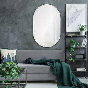Webster mirror with an oval shape and black frame, mounted on a textured gray wall above a gray sofa with a green blanket.