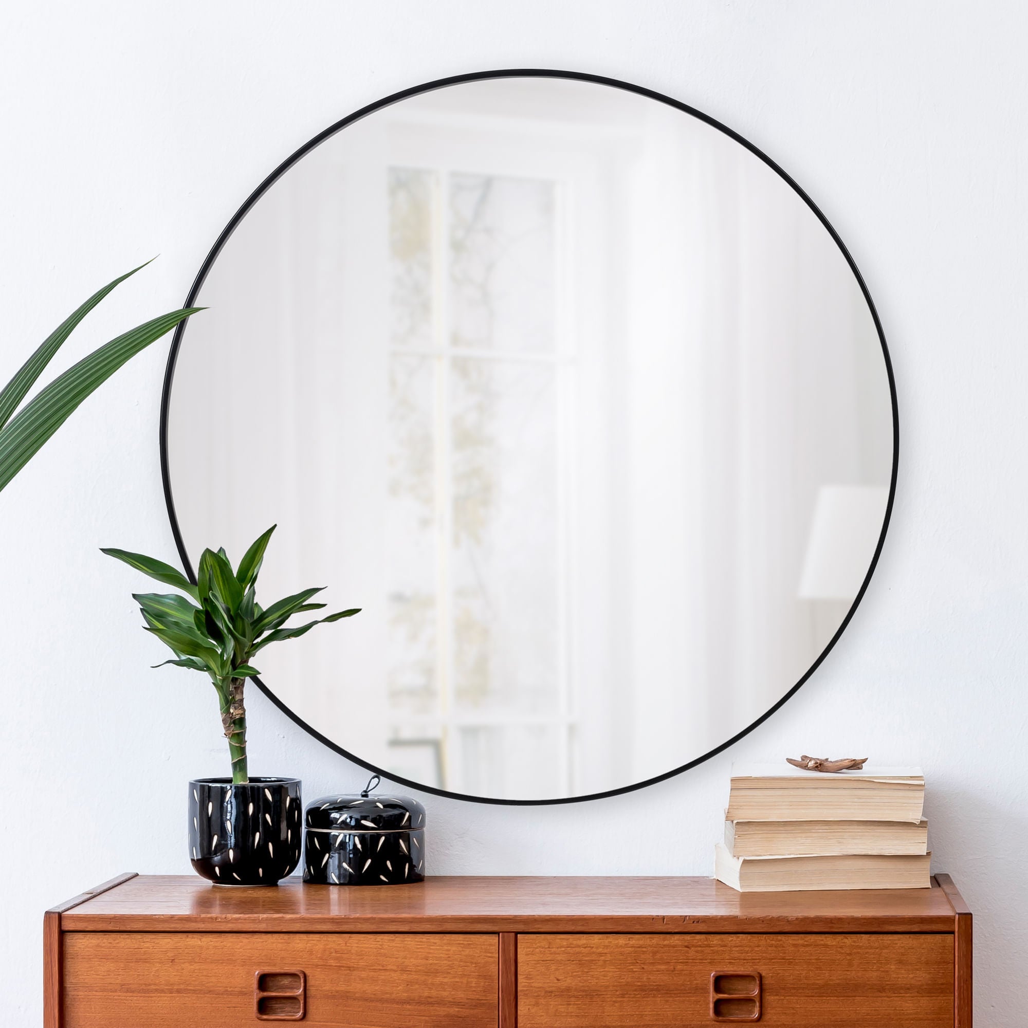 Cori round mirror with a black frame, displayed above a wooden cabinet with plants and books.