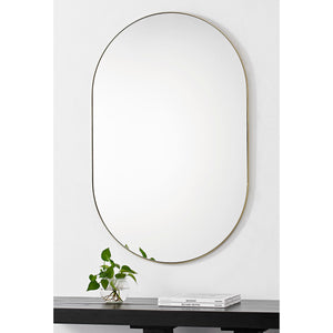 Beckah mirror with an oval shape and thin gold frame, mounted on a white wall above a black table with a plant.