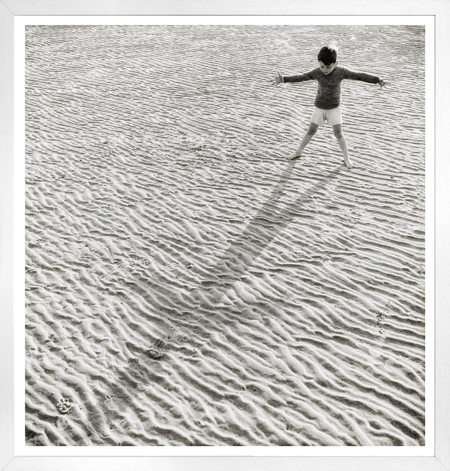 His Shadow, a monochrome wall art featuring a child playing on textured sand, casting a long shadow.