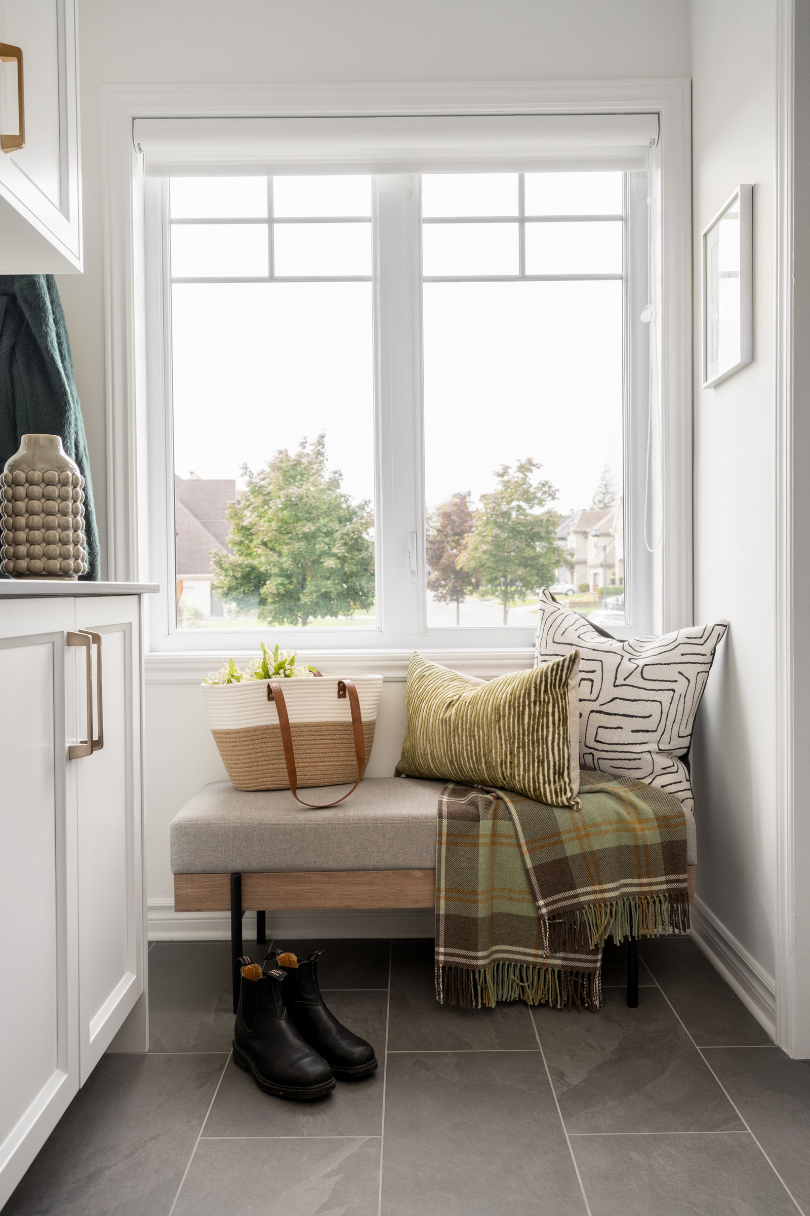 Styled mudroom nook with an upholstered bench, plaid throw, and woven tote beside a sunny window.