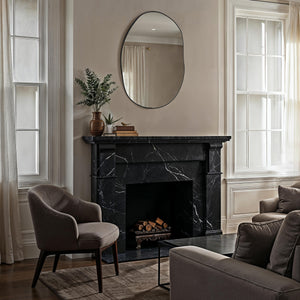 Messini mirror with an oval shape and black frame, hanging above a black marble fireplace in a cozy living room setting.