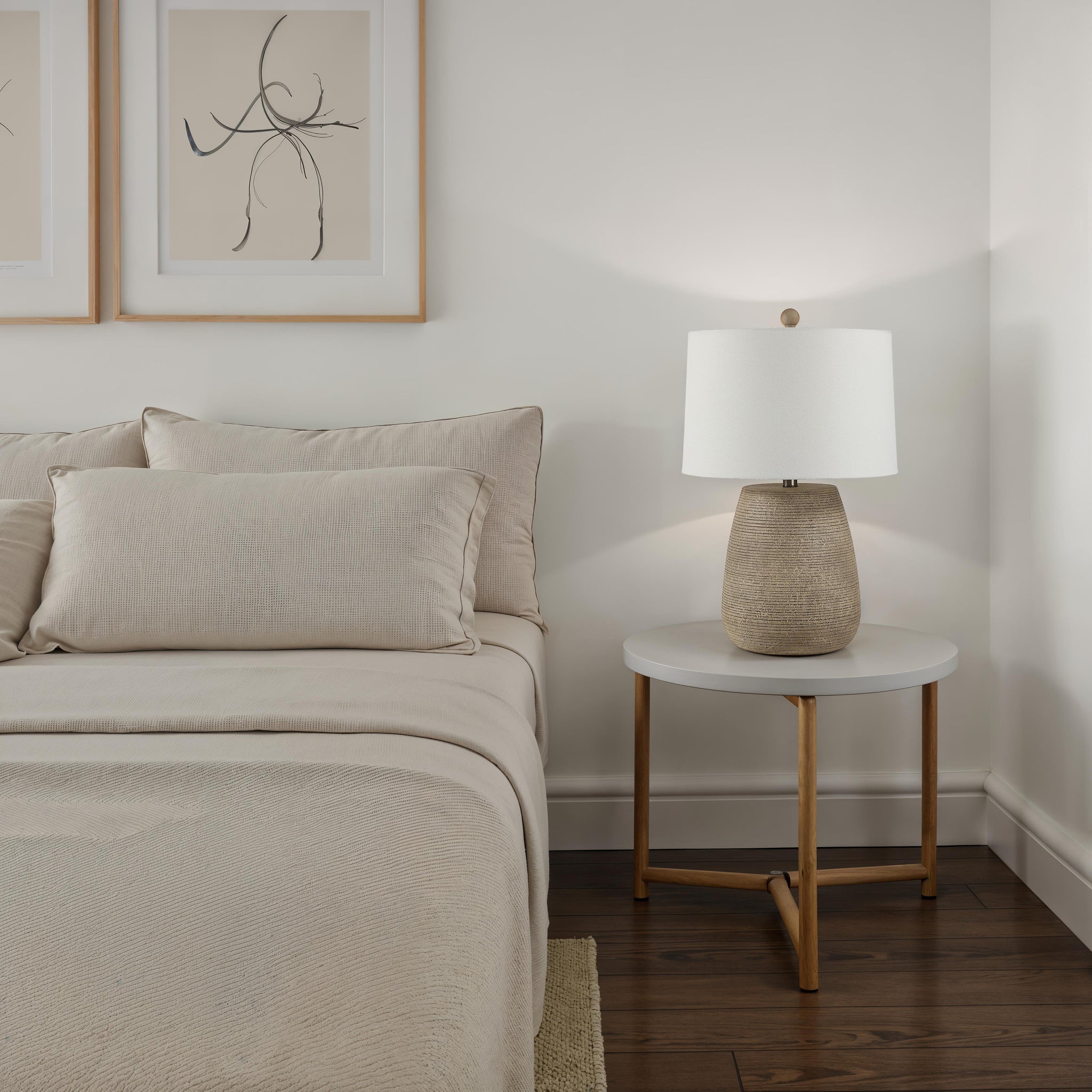 Optima table lamp on a bedside table next to a bed with beige linens and pillows.