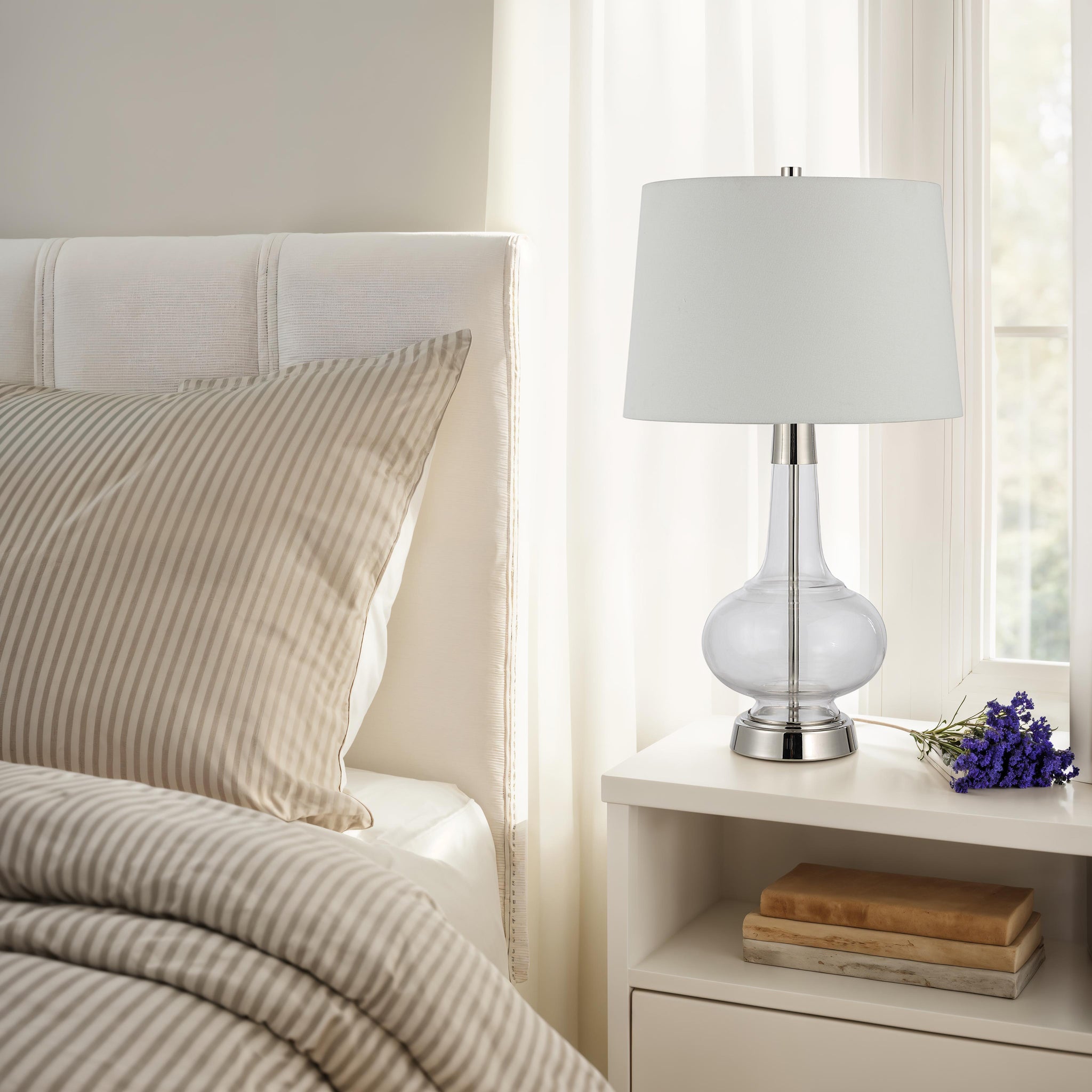 Lya Table Lamp on a nightstand beside a bed with striped bedding and a vase of flowers.