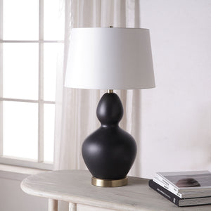 Kamilia Table Lamp placed on a table beside books, featuring a black ceramic body and white shade.