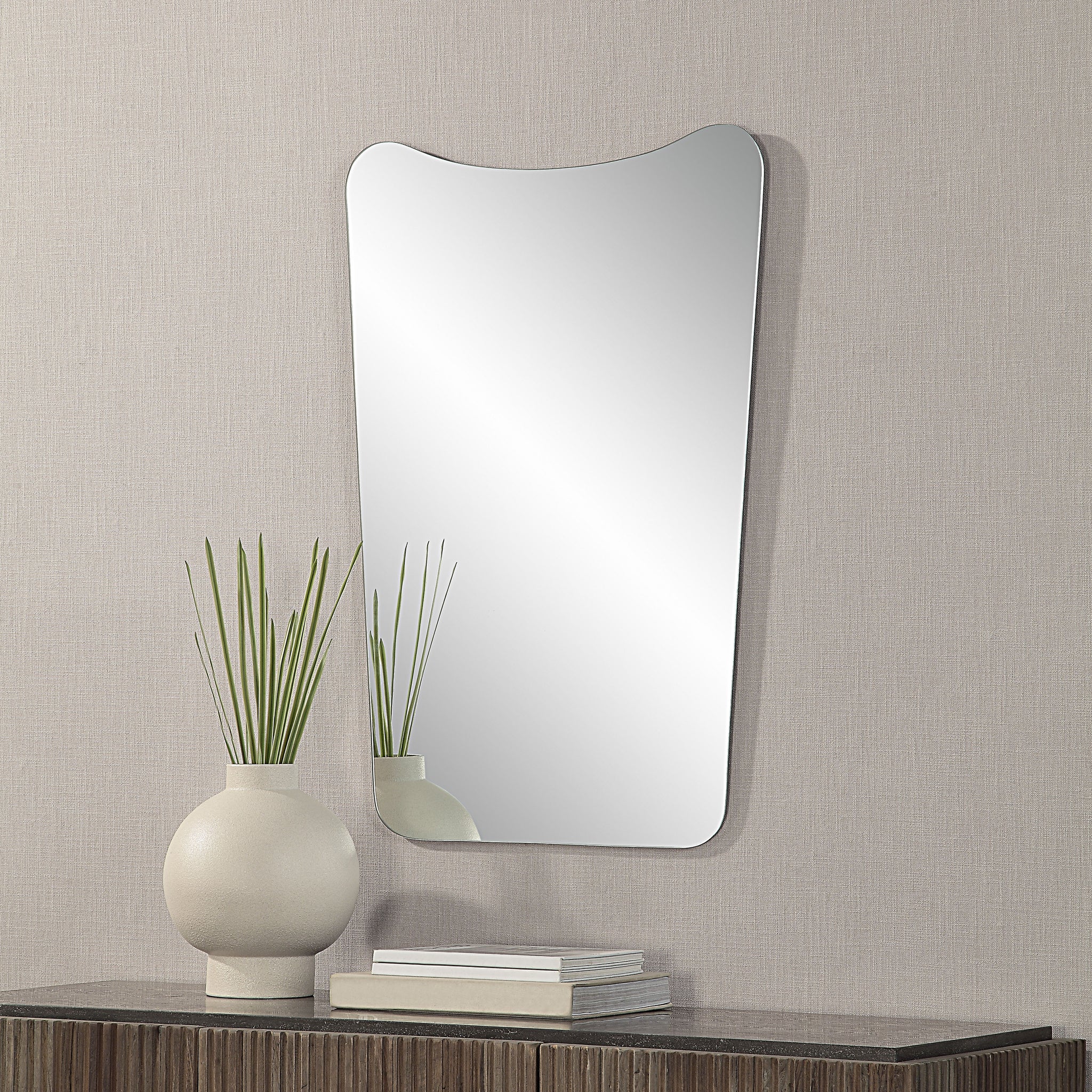 Selene Wall Mirror mounted on a textured wall, beside a vase and stacked books.