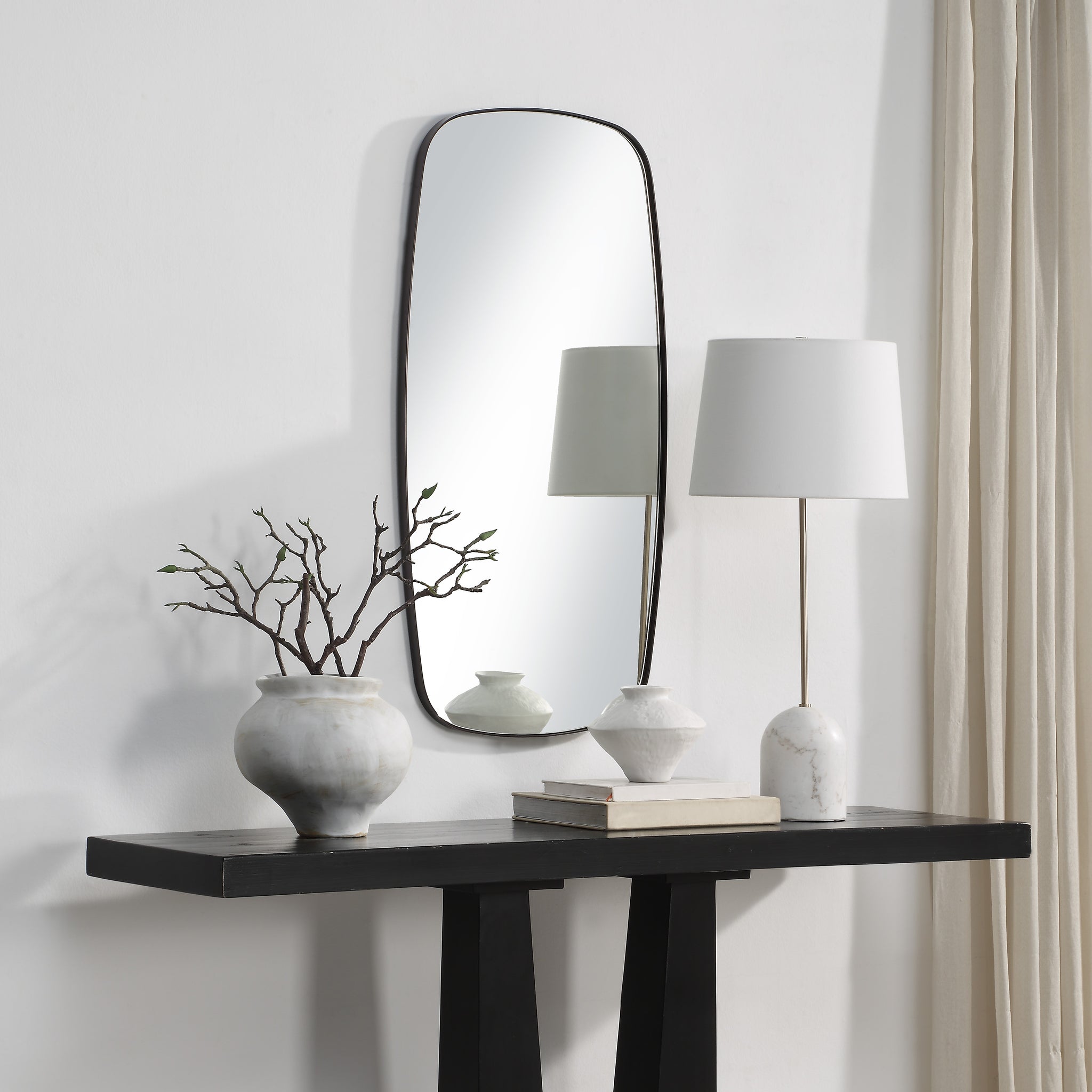 Nicoya Mirror displayed above a black console table with decorative vases and a lamp.